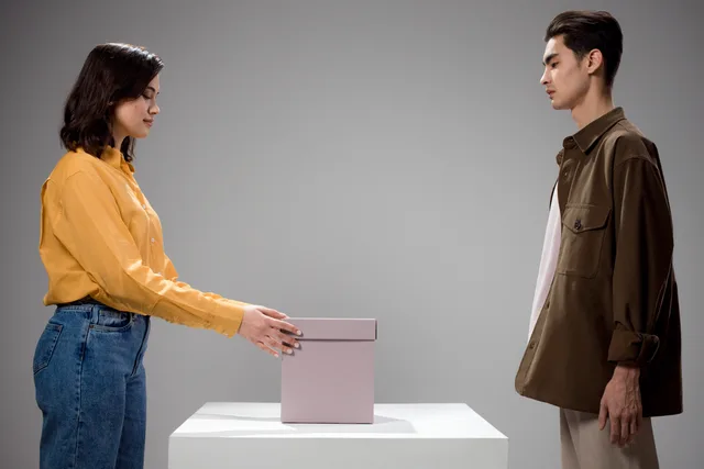 A man and woman standing opposite each other in a studio with a gift box in between. Returhantering enligt distansavtalslagen för e-handel